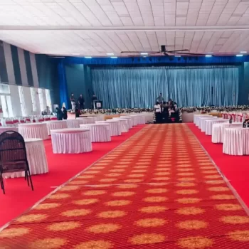 Large indoor event layout in Hyderabad by HKGN Tent House & Decoration, with round tables in white covers neatly placed on a bright red carpet and a stage at the back. A good example of the refined tent hire services we offer in the city.