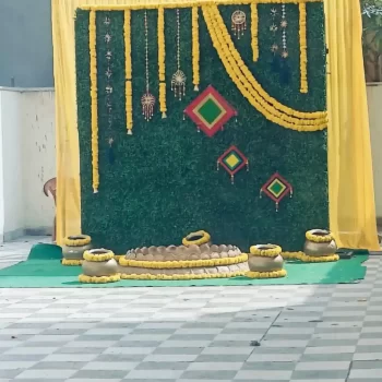 Decorated space arranged by HKGN Tent House & Decoration in Hyderabad, with a rich green backdrop hung with yellow marigold garlands and traditional Indian décor pieces. The tiled floor holds artistically placed decorative pots. A fine example of the tent and décor rental services HKGN Tent House & Decoration offers in Hyderabad.